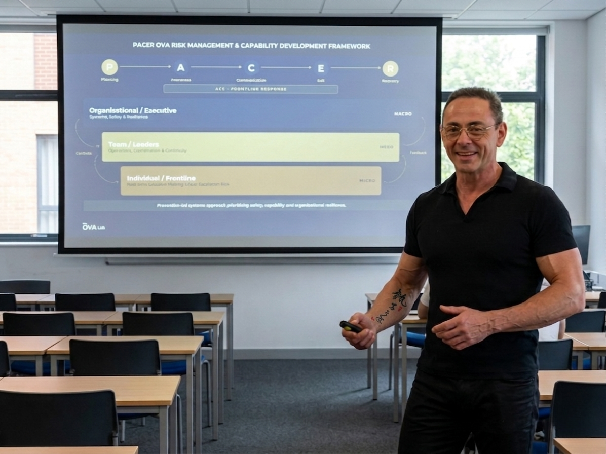 Smiling man in a black polo presents beside a projected slide in a classroom with desks and chairs ahead of him.